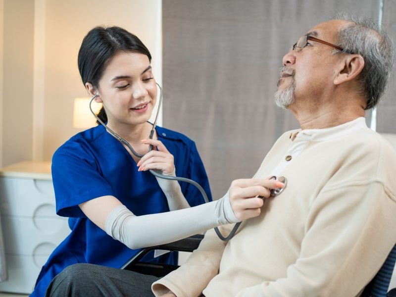 Asian nurse at nursing home taking care of senior man on wheelchair. Young woman therapist doctor measure heart rate by stethoscope on elderly older disabled patient. Medical insurance service concept