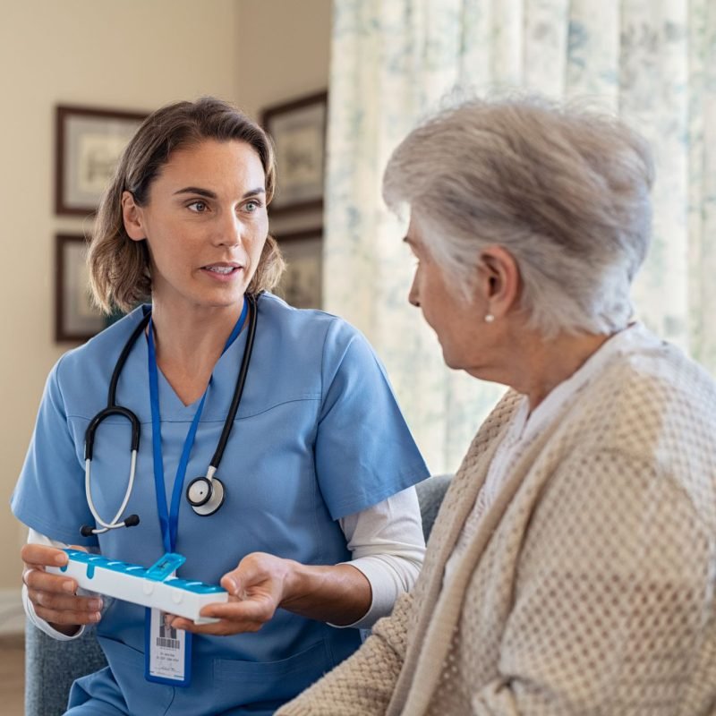 Young nurse explaining medicine dosage to old patient in care facility centre while holding weekly medicine dispenser. Professional caregiver holding pill organizer box giving medicine tablet to elderly woman. Nurse explaining medicine daily dosage for senior woman at care centre with copy space.