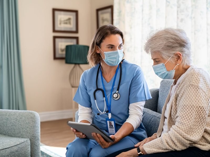 Young nurse and senior woman going through medical record on digital tablet during home visit and wearing face mask. Doctor wearing protective face mask during covid pandemic and showing medical reports to old woman at nursing home. Doctor and patient consultation at home during coronavirus and flu outbreak.